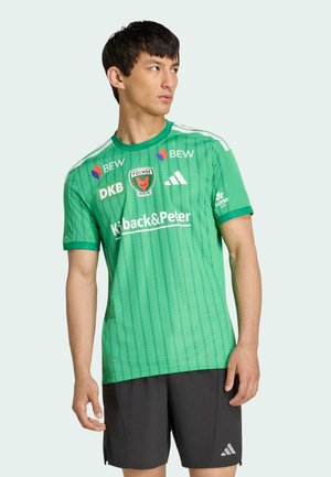 Mężczyzna ubrany w zieloną sportową koszulkę Fuchse Berlin z logo sponsorów oraz czarne spodenki Adidas, stojący i patrzący na bok.