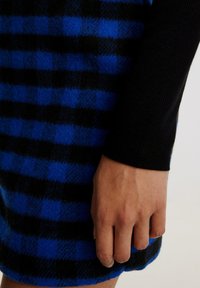Wollende rok met een blauw en zwart geruit patroon, gecombineerd met een zwart ribbelshirt met lange mouwen. Close-up van een hand die op het kledingstuk rust.