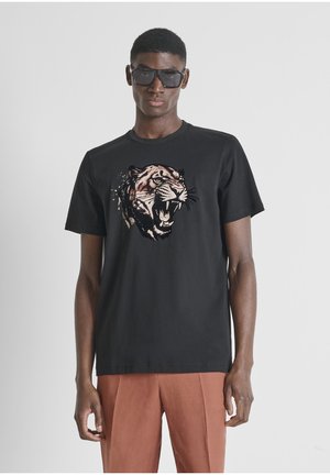 Joven con gafas de sol negras, camiseta negra con gráfico de tigre rugiente y pantalones color óxido, posando contra un fondo claro y liso.