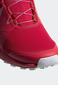 Chaussure de sport rouge Adidas avec tige en mesh, lacets argentés et semelle en caoutchouc blanche avec logo Adidas sur l’avant.