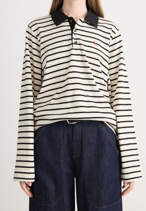 Kobieta ubrana w białą koszulkę polo z długim rękawem, z czarnymi poziomymi paskami i czarnym kołnierzem, w połączeniu z ciemnoniebieskimi jeansami.