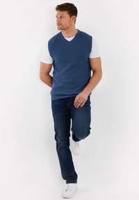 Blauwe gebreide mouwloze trui over een wit T-shirt, gecombineerd met donkerblauwe jeans en witte sneakers, wat een gelaagde casual uitstraling creëert.