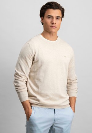 Hellbeige Rundhalsausschnitt-Pullover in einem Strickmuster, mit langen Ärmeln und gerippten Bündchen. Über hellblauen Hosen getragen, minimales Logodetail sichtbar.