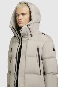 Beige pufferjakke med høj krave og stor hætte. Har lynlåslommer, sorte detaljer, quiltet tekstur og justerbare snore.