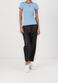 Osoba ubrana w dopasowaną jasnoniebieską koszulę z krótkim rękawem, luźne czarne skórzane spodnie oraz białe trampki, stojąca na jednolitym tle.