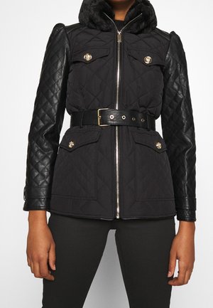 Frau trägt eine schwarze gesteppte Jacke mit Kunstlederärmeln, goldenen Knopfakzenten, Frontreißverschluss, tailliertem Gürtel und Fellkragen.