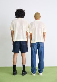 Kremfarvede kortermede strikkede skjorter med paisley-mønstre, parret med mørke shorts og løse denimjeans. Sorte og hvide sneakers bæres.