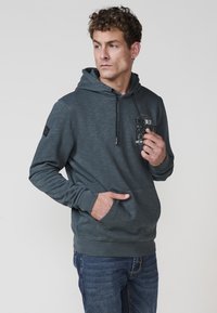 Sötétszürke kapucnis pulóver texturált felszíni kidolgozással, állítható húzózsinórral, elülső zsebbel, valamint logós részletezéssel a mellkason és a karon.