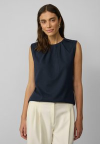 Mouwloze marineblauwe blouse met een zachte textuur, ronde halslijn en subtiele plooi-details aan de voorkant, gecombineerd met lichtgekleurde broeken.