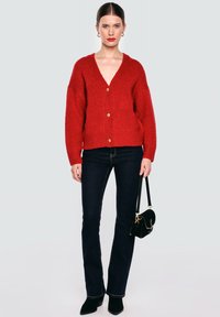 Červený pletený cardigan s výstřihem do V, třemi zlatými knoflíky, žebrovanými manžetami a lemem. Spárovaný s tmavými džíny a malou černou kabelkou.