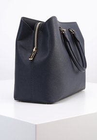 Borsa a tracolla in tessuto blu navy con accessori dorati, chiusura con zip e due manici superiori; forma ampia e strutturata con una finitura liscia e lucida.