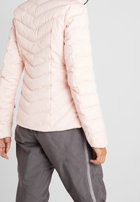 Světle růžová prošívaná péřová bunda s kosočtverečkovým šitím, kapsami na zip a přiléhavým střihem, doplněná šedými kalhotami. Měkká textura a teplý materiál.
