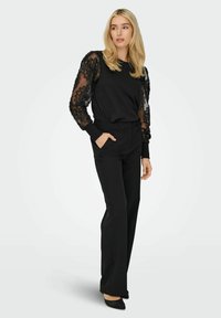 Top nero con maniche trasparenti a fantasia floreale, abbinato a pantaloni neri sartoriali. L'outfit presenta una vestibilità rilassata e una silhouette moderna.