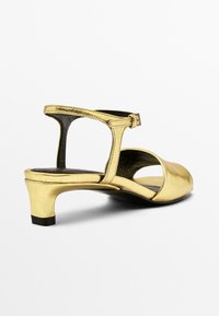 Des sandales à talons métalliques dorées avec bout rond, côtés ouverts, un design à sangle en T et un petit talon en bloc avec une finition texturée.