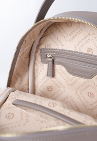 Mochila de cuero gris con un interior crema, cuenta con un bolsillo con cremallera y un patrón de logo en relieve en el forro. Cremallera dorada.