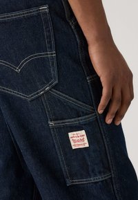 Calças de ganga azul-escuro com múltiplos bolsos e costura branca visível, apresentando uma mão repousada perto de uma pequena etiqueta da Levi Strauss & Co.