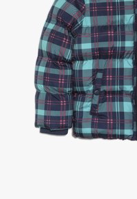 Veste matelassée bleu marine avec un motif à carreaux turquoise et rose. Elle présente une fermeture à boutons sur le devant et des poignets en tricot.