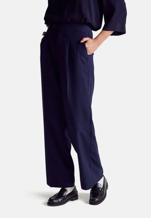 Pantaloni navy a gamba larga con pieghe frontali, tasche laterali, una texture liscia e una vestibilità su misura, abbinati a mocassini neri e calze grigie.