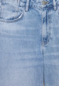 Jean en denim bleu clair avec poches avant, fermeture par bouton métallique, passants de ceinture et détails de couture visibles.