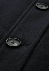 Czarny wełniany płaszcz z detalami, w tym dwiema dużymi okrągłymi guzikami oraz widocznym przeszyciem wzdłuż patki, ukazujący miękką, teksturowaną tkaninę.