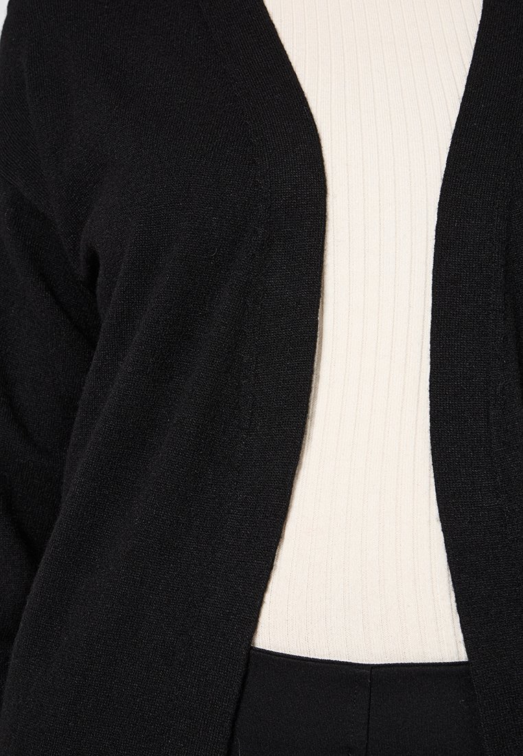 Czarny sweter z dzianiny z otwartym przodem, charakteryzujący się miękką teksturą. Nosi się go na kremowej bluzce z prążkami, co podkreśla kontrastujące kolory i warstwy.