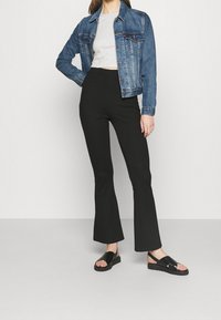 Chaqueta de mezclilla con corte clásico, de color azul; combinada con pantalones de campana negros de cintura alta y sandalias negras de tiras. Conjunto sencillo y moderno.