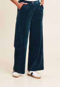 Pantalons en velours côtelé à jambes larges de couleur bleu sarcelle profonde avec une texture douce, dotés de plis avant, de poches latérales et d'une fermeture à bouton discrète.