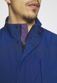 Blå jacka med hög krage, med lila innerfoder, svart dragkedja och tryckknappar. Slät textur med ett subtilt mönster.