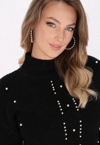 Maglione nero a coste con decorazioni in perle disposte in un motivo verticale. Collo alto e maniche lunghe. Il modello ha capelli lunghi e ondulati.