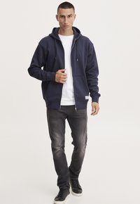 Marineblå zip-up hettegenser laget av bomullsblend, med en frontlomme, snorhette og ribbestrikkede mansjetter, sammen med grå jeans og svarte joggesko.