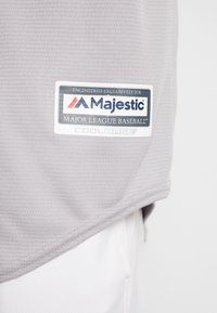 Jersey de béisbol gris hecho de tejido texturizado, con una etiqueta de logo en blanco y azul de Majestic, con el texto "COOLBASE" y "MAJOR LEAGUE BASEBALL".