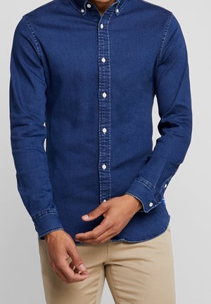 Mann trägt ein eng anliegendes dunkelblaues Jeanshemd mit Knöpfen und beige Hose, richtet die Manschette eines Ärmels.