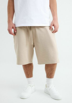 Mand iført beige shorts med snøre, hvide mellemlange sokker, hvide sneakers og en hvid t-shirt, stående på en hvid baggrund.