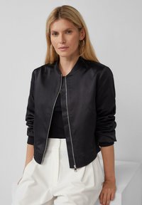 Giubbotto bomber nero con finitura liscia e lucida, chiusura a zip frontale e polsini a coste. Indossato sopra una maglietta nera e pantaloni bianchi. Design minimalista.