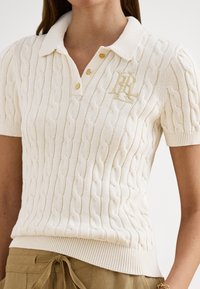 Kvinna bär en vit kortärmad polotröja i kabelstickning med guldfärgade knappar och en broderad logotyp på bröstet, kombinerad med beige byxor.