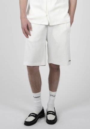 Mężczyzna w białych teksturowanych krótkich spodenkach, białej koszuli, białych markowych skarpetkach i czarno-białych mokasynach stoi na jednolitym tle.
