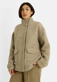 Mujer vestida con una chaqueta de lana beige con cuello alto y bolsillos con solapa, combinada con pantalones beige a juego, de pie contra un fondo liso.