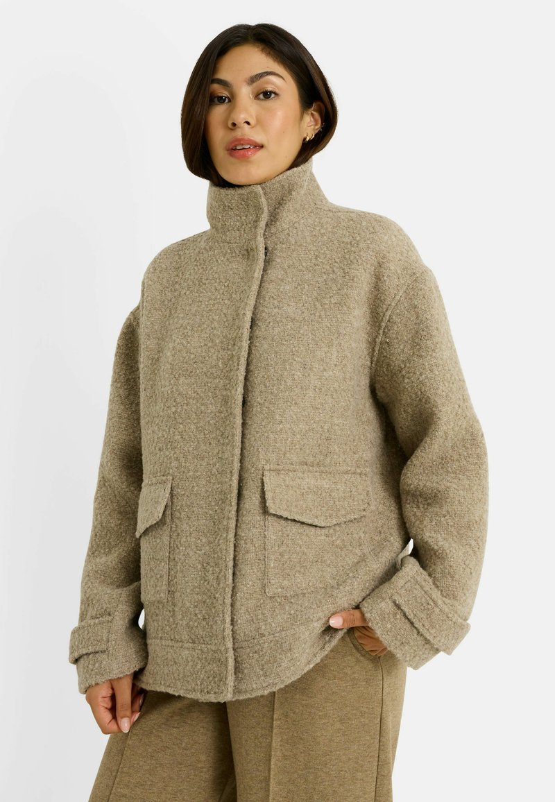 Mujer vestida con una chaqueta de lana beige con cuello alto y bolsillos con solapa, combinada con pantalones beige a juego, de pie contra un fondo liso.