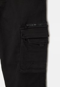 Pantalon cargo noir en tissu lisse avec plusieurs poches, une poche zippée et des accents de couture robustes.