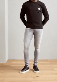 Svart sweatshirt med ett litet emblem, grå slim-fit jeans och svarta sneakers med vita sulor, placerade på ett trägolv mot en ljus vägg.