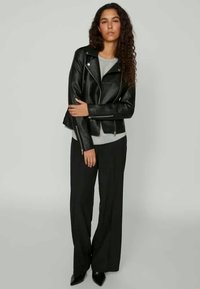 Jaqueta de motociclista em couro preto com um design ajustado, acessórios em prata e bolsos com fecho de correr, usada sobre uma blusa cinza e calças largas pretas.
