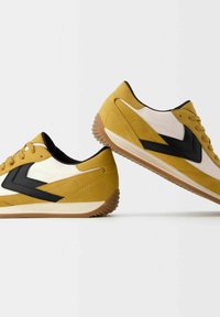 Gula och vita sportskor med svarta detaljer. Tillverkade av mocka och nät, med gummisula och kantiga designdetaljer.