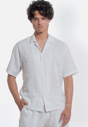 Giovane uomo con capelli ricci che indossa una camicia bianca a maniche corte abbottonata e pantaloni bianchi coordinati, in piedi con una mano in tasca.