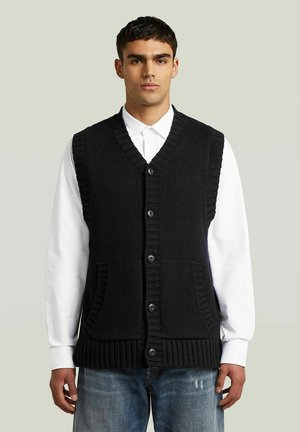 Junger Mann trägt eine schwarze, gestrickte, knopfgeschlossene Weste über einem weißen Hemd und blauen Jeans, steht vor einem einfarbigen hellen Hintergrund.