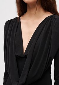 Czarna sukienka z głębokim dekoltem w kształcie litery V, drapowanym materiałem wokół biustu oraz marszczeniem na ramionach, ukazująca gładką fakturę.