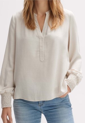 Kobieta ubrana w luźną, kremową bluzkę z długim rękawem z marszczonymi mankietami oraz jasnoniebieskie dżinsy, stojąca z jedną ręką w kieszeni.