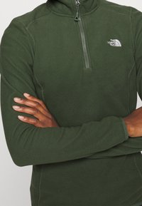 Pullover in pile verde scuro con colletto alto, zip frontale a metà e logo The North Face sul petto. Tessuto morbido, maniche lunghe aderenti.