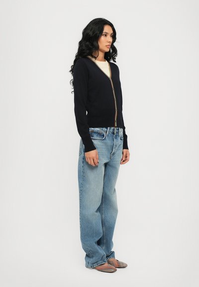 Cardigan blu navy con maniche lunghe e una vivace patta di bottoni, abbinato a jeans in denim chiaro a vestibilità ampia e scarpe slip-on a righe.