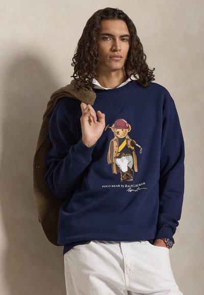 Moški z dolgimi kodrastimi lasmi, ki nosi mornarsko modro majico s potiskom Polo medveda in bele hlače, drži rjavo jakno čez ramo.