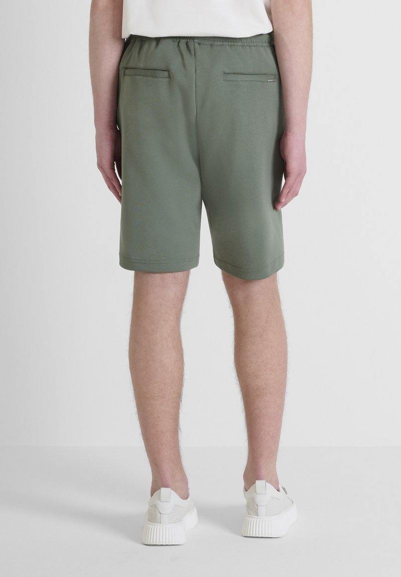 Shorts verts avec une ceinture élastique, deux poches arrière et une texture lisse. Associés à des baskets blanches, vus de l'arrière.